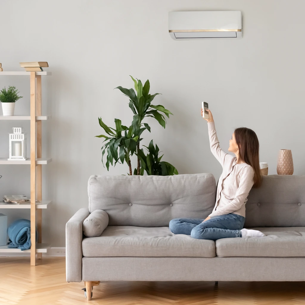 Frau sitzt auf einem grauen Sofa und steuert eine Klimaanlage an der Wand mit einer Fernbedienung.