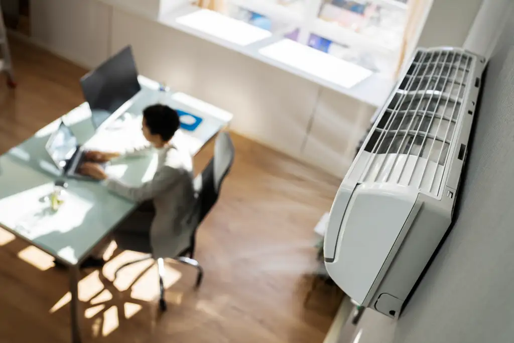 Wandklimagerät in einem modernen Büro bläst kühle Luft, während eine Person am Schreibtisch mit einem Laptop arbeitet.