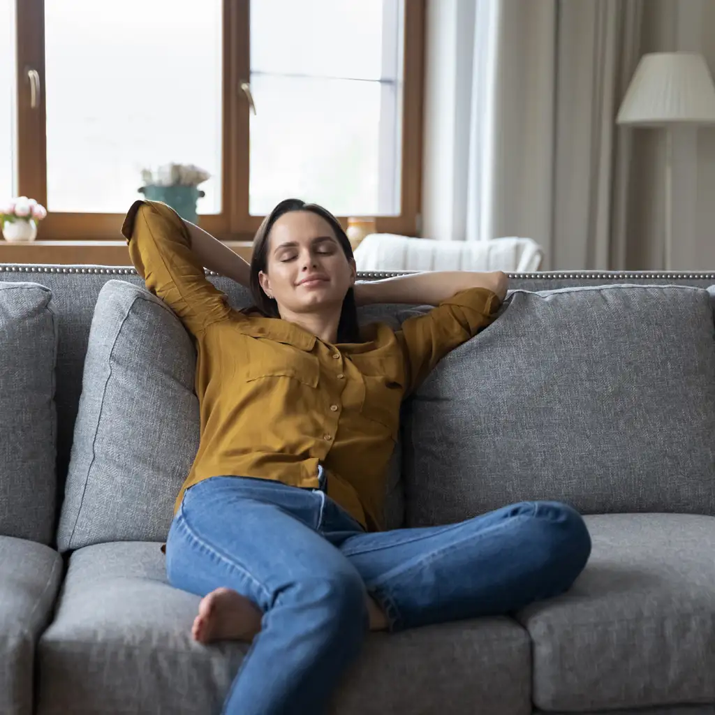 Bestes Raumklima: Eine Frau sitzt entspannt auf einem grauen Sofa mit geschlossenen Augen und genießt die angenehme Atmosphäre in ihrem Wohnzimmer.